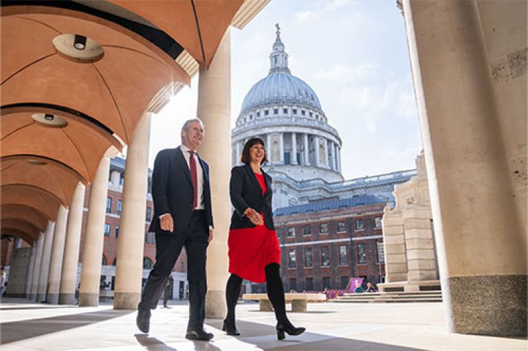 Prime Minister Keir Starmer and Chancellor Rachel Reeves visit the London Stock Exchange. They are smartly dressed and walk. Behind them are columns, and further in the distance, the rounded dome of St Paul's Cathedral in London.