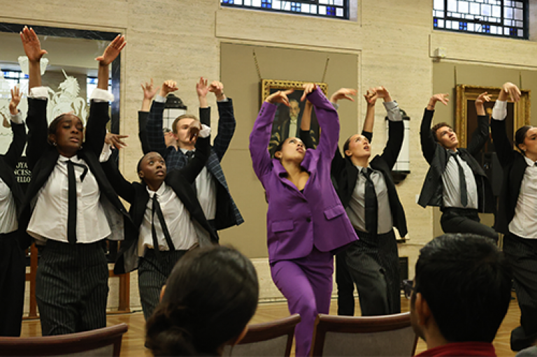 A person in a purple suit dances surrounded by performers in black suit