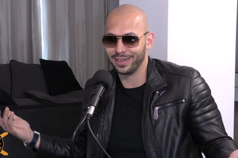 An image of Andrew Tate. He is bald with a beard and wears dark sunglasses. He wears a black leather moto jacket and smiles as he speaks into a microphone. Behind him is a white wall, a grey curtain and a black couch.