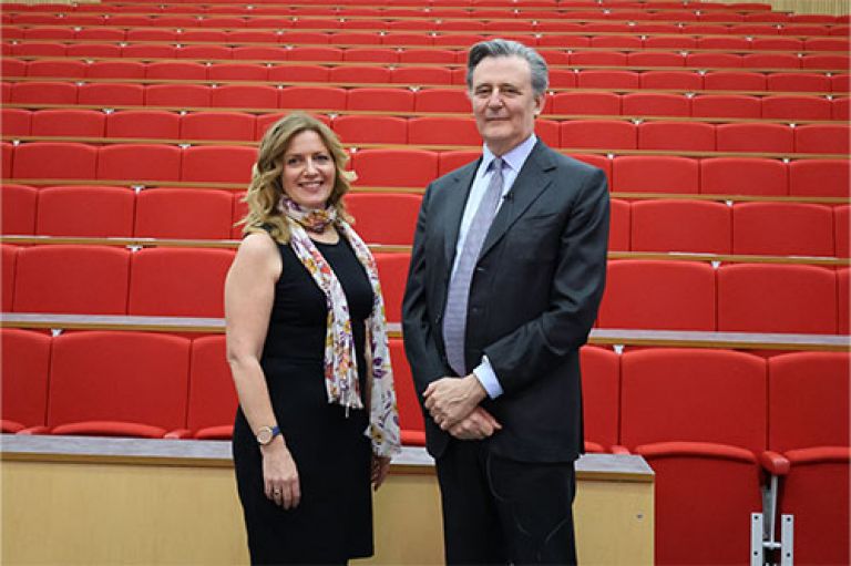 An image of Dr Glenda Cooper, wearing a dress and smiling to camera, standing next to John Micklethwait, wearing a suit and smiling to camera. They are standing in front of rows of empty red seats.