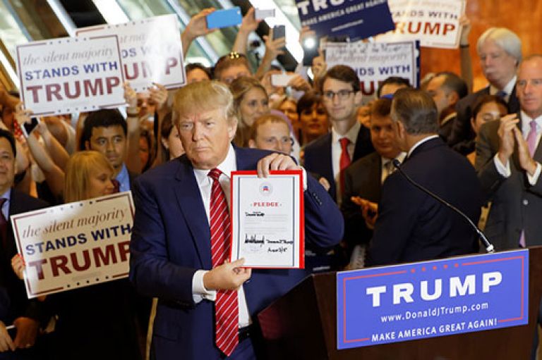 Donald Trump holds a pledge to camera at a campaign rally. All around him are people wearing suits and holding signs that read 